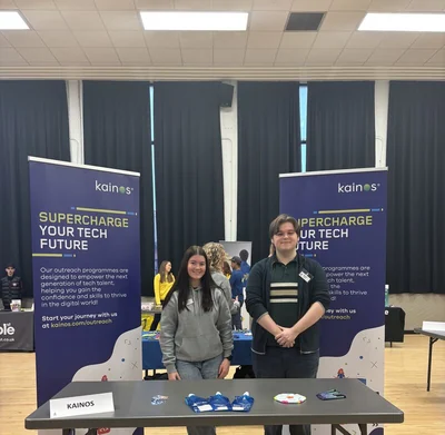 Two people at a Kainos outreach table between purple banners at Ashfield Boys' High School