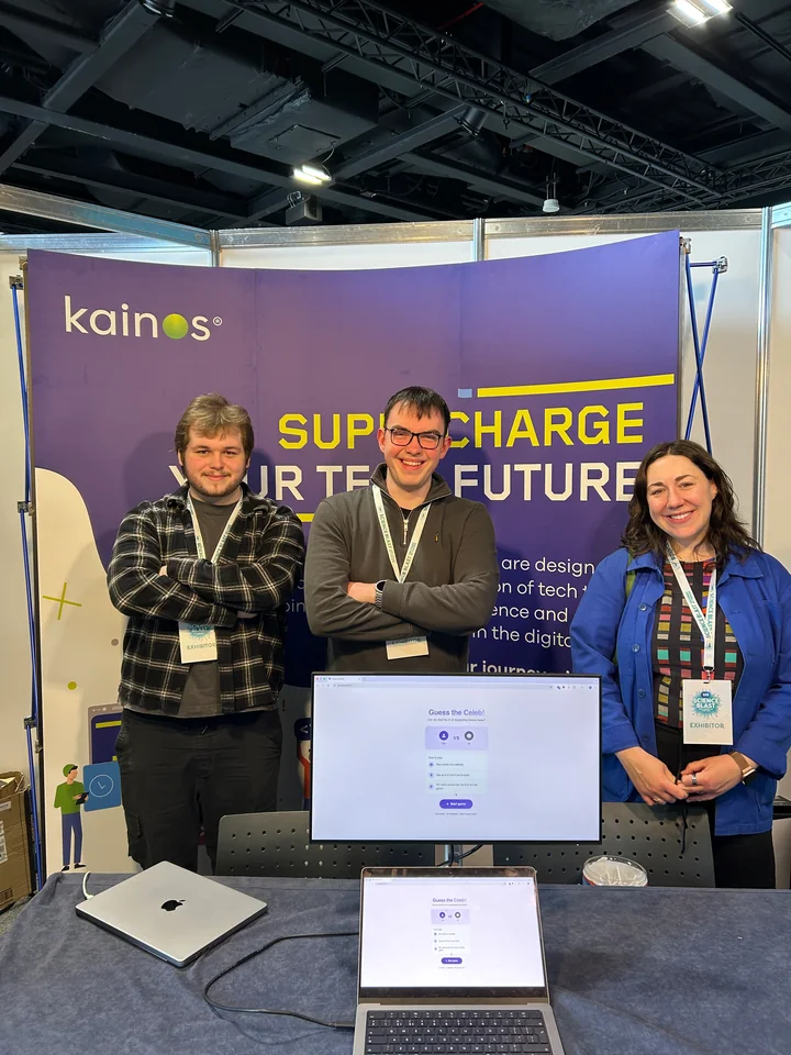 Three people at a Kainos stand with a game displayed on a monitor